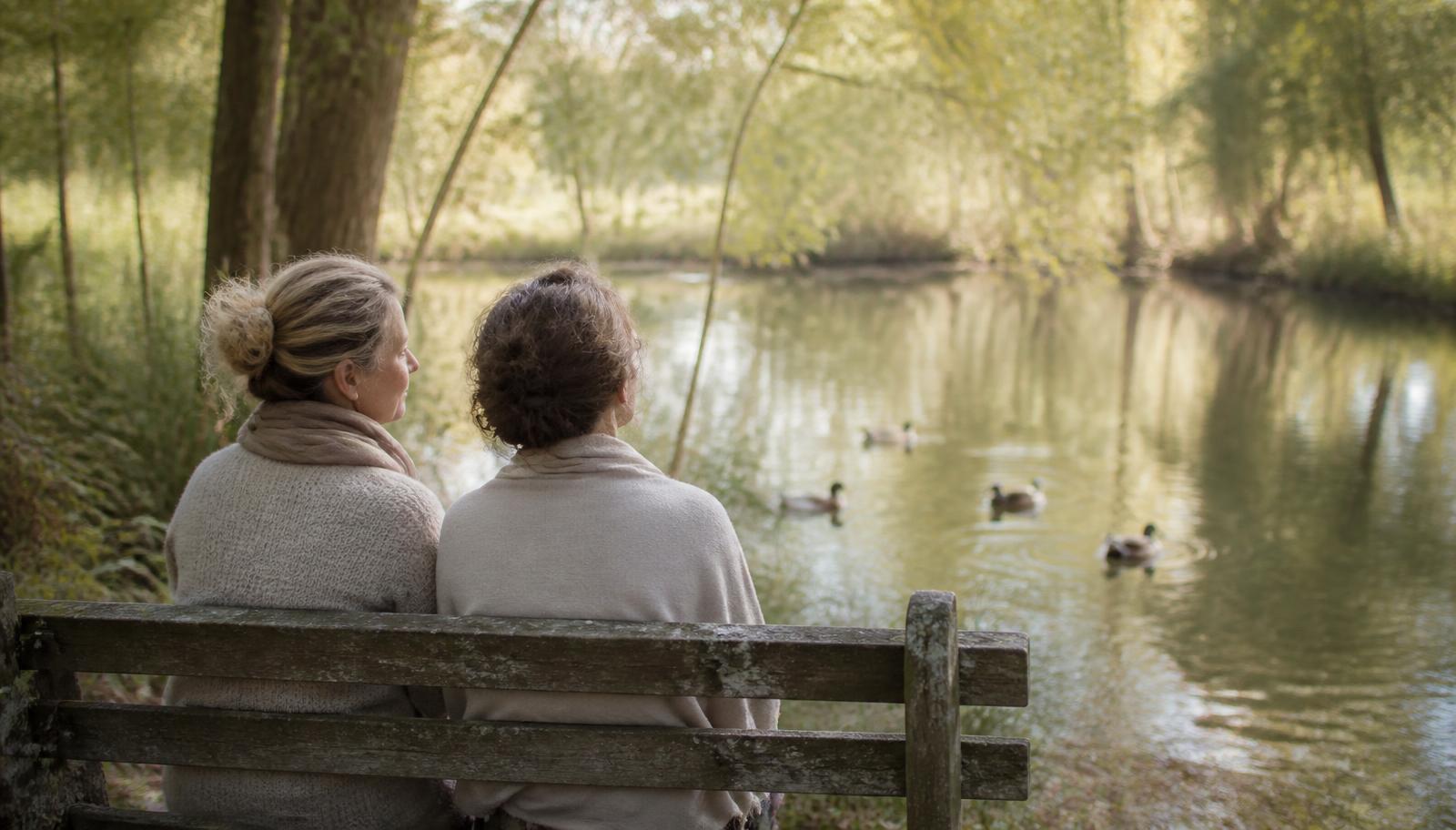 Two women sitting on a bench beside a quiet pond, watching ducks drift on the water